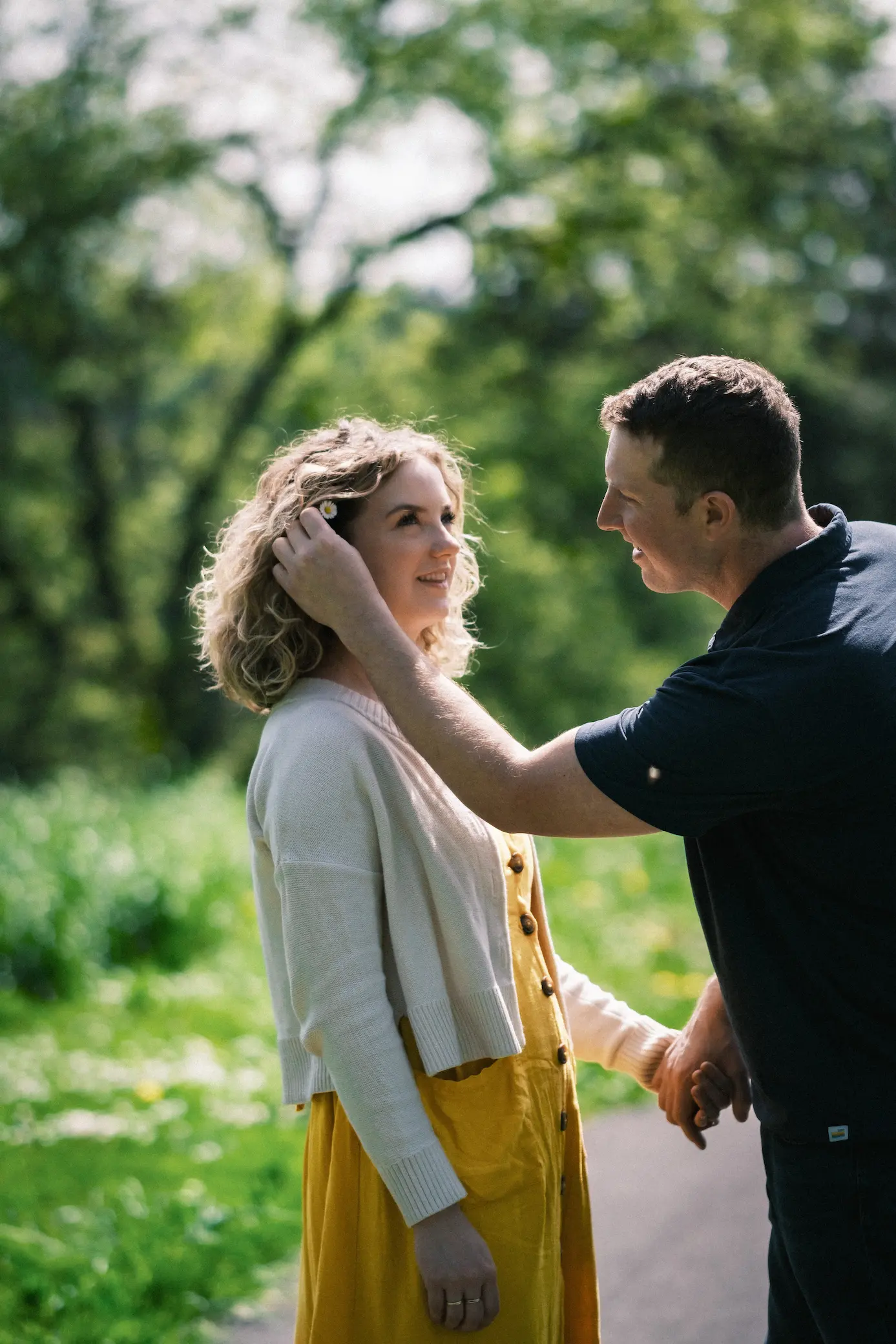 Couple in the park with one person placing a daisy behind the others ear