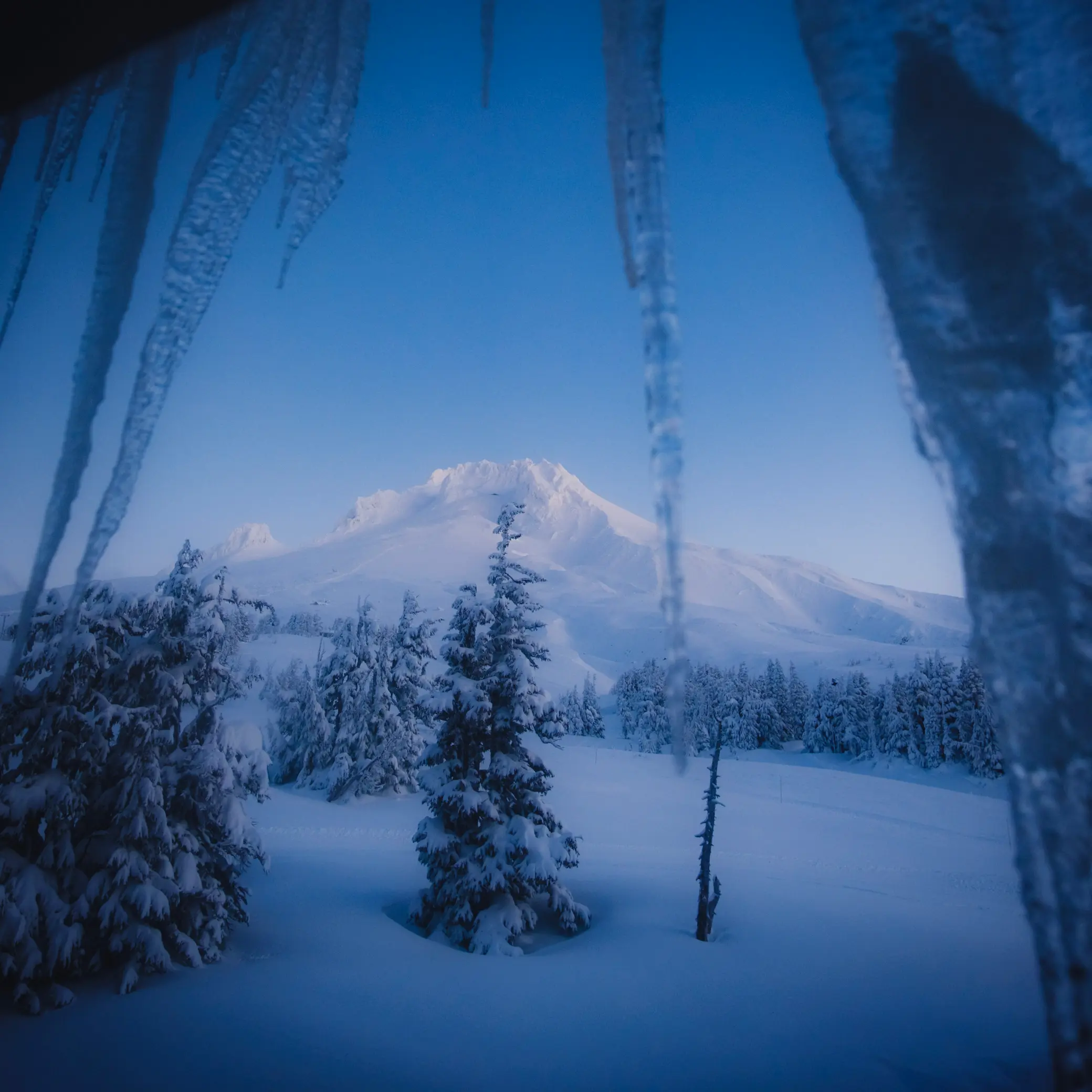 Icicles hang from the window as a mountain in snow at dusk is in frame