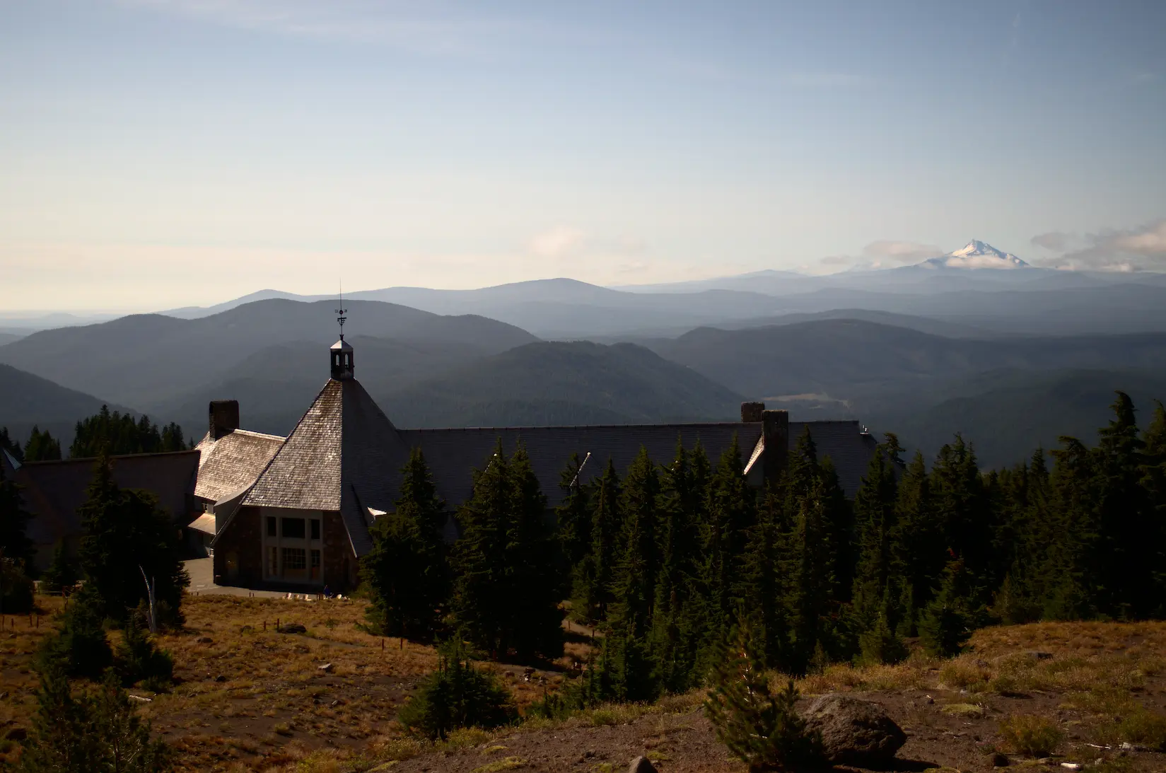 A lodge with some trees surrounding it overlooks a valley with another mountain in the background